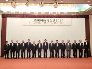 三陸復興防災会議2023の横断幕が貼られたステージ前にスーツ姿の男性が15名並んでいる写真