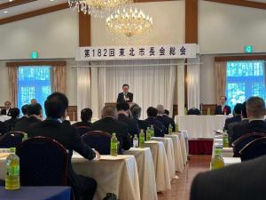 演台の前でマイクを持って話す男性と椅子に座って演台の方を向く参加者たちの写真