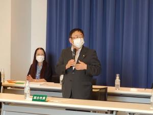 「宮古市長」と書かれた緑色のネームプレートが置かれた机の前に立ち、右手にマイクを持って話をしている様子の市長と、後方で席に着き話を聞いている様子の女性を写した写真
