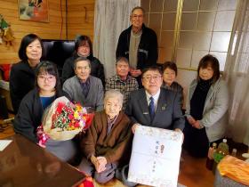 ご自宅の居間で、市長が市からの記念品を持ち、ご家族の1人が花束を持って、上山ウメチさんを中心にして8人のご家族が集合して撮影した写真