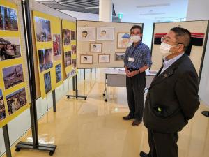 並べられたボードに貼られた写真や絵が展示された会場で、関係者の男性1名と市長が鑑賞している様子の写真