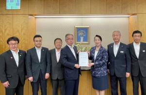 壁に瓶に生けられた花の絵画が設置された壁をバックに、中央に総務省の尾身朝子副大臣と市長会の代表が一緒に要望書を持って中央に立ち、市長を含む5名の関係者が横一列に並んでいる写真