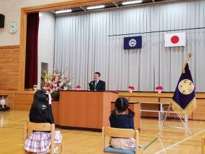 体育館のステージ下に置かれた演台で、市長が座っている新1年生に向けて祝辞を述べている写真