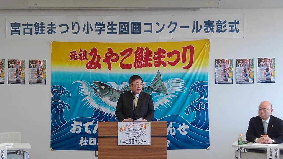 「宮古鮭まつり小学生図画コンクール表彰式」と書かれた横幕と「元祖みやこ鮭まつり」と書かれた鮭が荒波にもまれたイラストが描かれた旗が掲示された会場で、檀上に立つ市長が挨拶を行っている表彰式の様子の写真