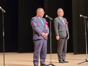 舞台上に置かれたスタンドマイクの前で、赤い胸章を着け両手を体の前で組み話をする市長と、その横で直立している男性を写した写真