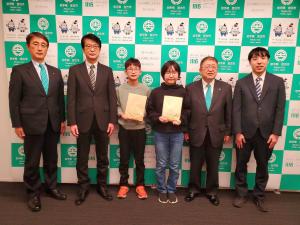 宮古市のバックパネルをバックに高浜小学校児童2名が能登半島地震災害義援金の箱を持って市長と関係者3名にはさまれて記念撮影をしている写真