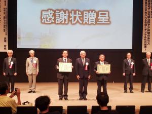 スクリーンに感謝状贈呈の文字が映し出されたステージ上に賞状を持った市長と男性の間に胸章をつけた男性が立ち、ステージ後方に胸章をつけた男性が4名立っている写真