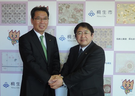 桐生市のバックパネルの前で両手で握手をする山本市長と群馬県桐生市の亀山豊文市長の記念写真