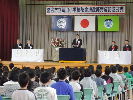 会場に並べられた椅子に座る小学生たちと、舞台上の演台のマイクに向かい話をする市長の写真