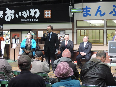 魚菜市場のお店の前で、マイクを持って立ちながら話をする市長と、後方で椅子に座る関係者の男性3名、観客の方々を写した写真