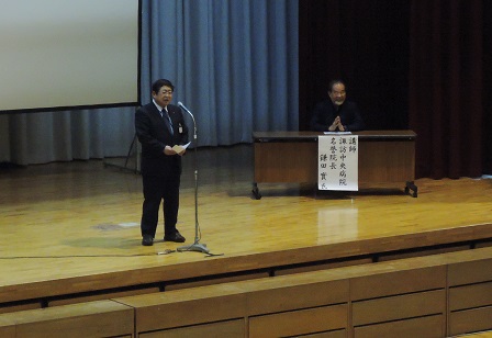 舞台上で、名前札が掛けられたテーブルの席に着く男性と、中央でスタンドマイクに向かい話をする市長の写真