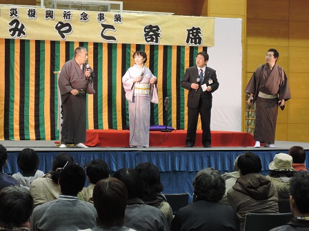「みやこ寄席」と書かれた横断幕が掲げられた舞台上で、スーツを着た市長、着物を着た2名の男性と1名の女性が立っている写真