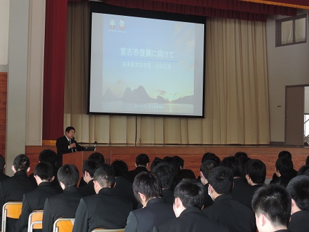 画面が映されたスクリーンの横の演台で、マイクを持ち講演する様子の市長と、会場に並べられた椅子に座る大勢の学生たちの写真