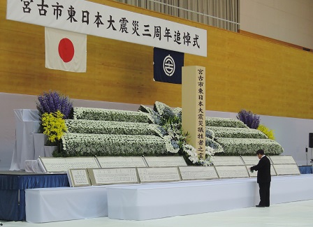 「宮古市東日本大震災三周年追悼式」と書かれた横断幕、日本国旗、宮古市の市町村旗が掲げられ、白色、黄色、紫色の花がたくさん飾られた献花台の前に立つ市長の写真