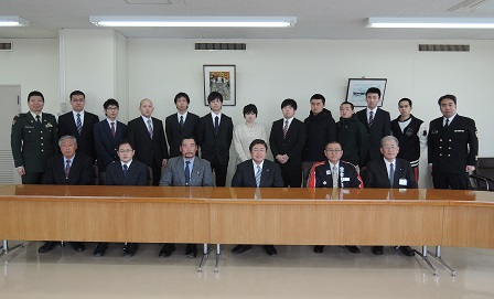 長机の後方で椅子に座る市長と5名の男性、その後ろで立って横一列に並ぶ13名の関係者の方々の記念写真