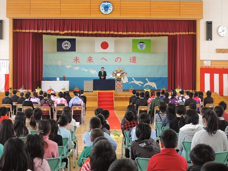 体育館に並べられた椅子に座る小学生たちと、舞台上の演台のマイクに向かい話をする市長の写真