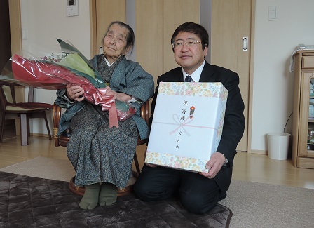 熨斗がついた記念品の箱を持ってしゃがむ市長と、椅子に座り花束を持つ女性の写真