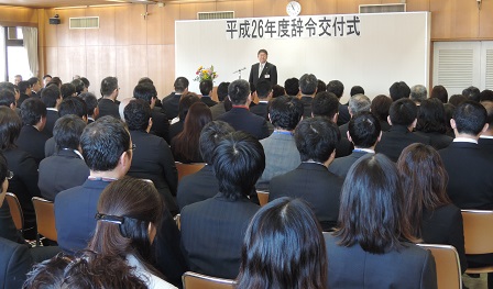 「平成26年度辞令交付式」と書かれた紙が貼られたボードの前に立ち話す市長と、並べられた椅子に座る大勢の職員たちの写真