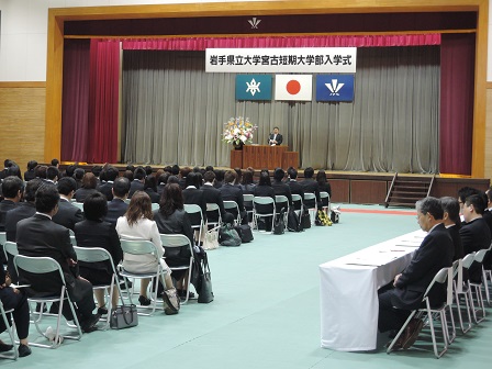 パイプ椅子に座る学生たちや保護者と、舞台上の演台のマイクに向かい話をする市長を写した写真
