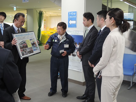 写真のパネルを持った男性と、パネルの方向に手を向ける市長、向かい側に立ち話を聞く様子の3名の関係者の写真