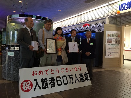 「おめでとうございます 祝 入館者60万人達成」と書かれたパネルの後ろに立つ、熨斗がついた記念品を持つ市長と男性、額縁を持つ男性、花束を持つ女性、両手をお腹の前で組む男性が並んで立つ記念写真
