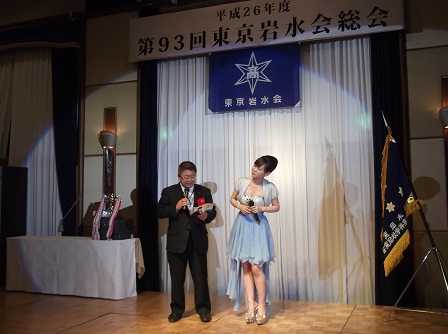「平成26年度第93回東京岩水総会」と書かれた横断幕が掛けられた舞台で、青色のドレスを着たみやさと奏さんと市長が立っている写真