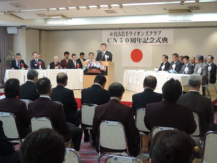 舞台上で左右に分かれて並べられた机の席に関係者が座り、中央で演台に置かれたマイクに向かい話をする市長の写真