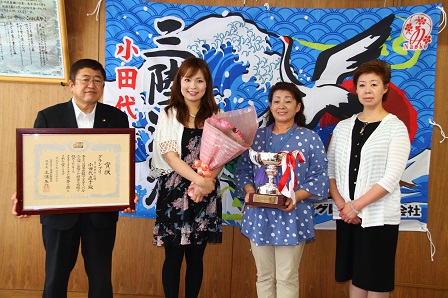 大漁旗の前で賞状の額縁を持つ市長、花束を持った女性、トロフィーを持った女性と両手をお腹の前で組む女性が横一列に並ぶ記念写真