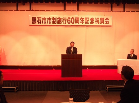 「黒石市市制施行60周年記念祝賀会」と書かれた横断幕が吊るされた舞台上で、演台に置かれたマイクに向かい話をする市長の写真