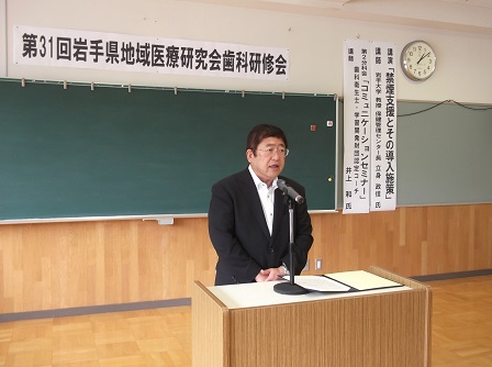 「第31回岩手県地域医療研究会歯科研修会」と書かれた紙が貼られ、黒板が設置された壁の前で、演台に置かれたマイクに向かい話をする市長の写真