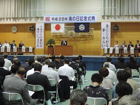 「祝 平成26年海の日記念式典」と書かれた横断幕、日本国旗、宮古市の市町村旗が吊るされた舞台上で、演台に向かい立つ市長と、会場に並べられたパイプ椅子に座り整列する関係者の方々の写真