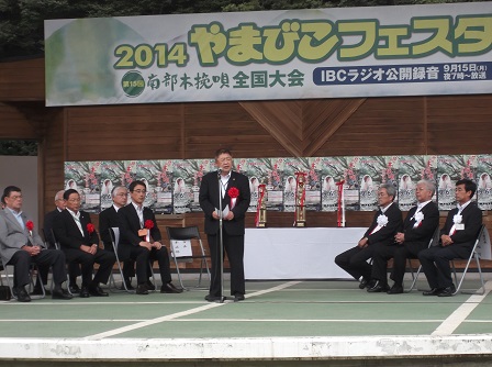 トロフィーが置かれた机の前で左右に分かれて並べられた椅子に座る関係者の男性たちと、中央でスタンドマイクに向かい話をする市長の写真