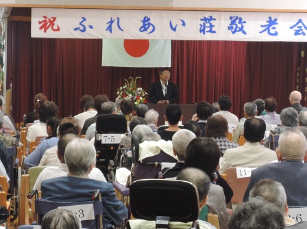 椅子に座る高齢者たちと、演台に置かれたマイクに向かい話をする市長の写真