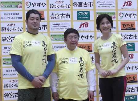 企業のロゴが載せられたバックパネルの前で、市長を中心に高田延彦さん、向井亜紀さんが並んでいる記念写真