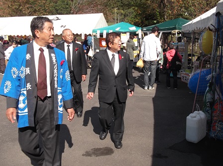 法被を着た男性とスーツを着た市長が、並んだテントを見ながら歩いている写真