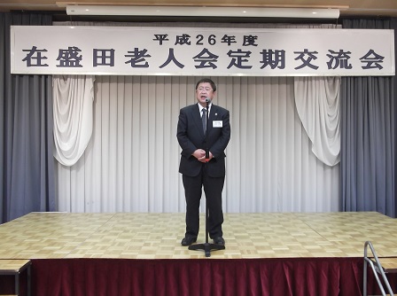 「平成26年度 在盛田老人会定期交流会」と書かれた横断幕が掲げられた舞台上で、スタンドマイクに向かい話をする市長の写真