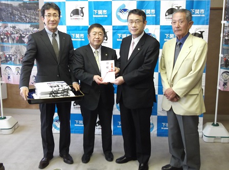 目録を一緒に持つ市長と男性、お盆を持つ男性、黄色いジャケットを着た男性が並んでいる記念写真