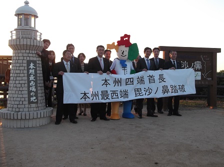 「本州四端首長 本州最西端 毘沙ノ鼻踏破」と書かれた横断幕をもつ市長と関係者の男性、よんたんの記念写真