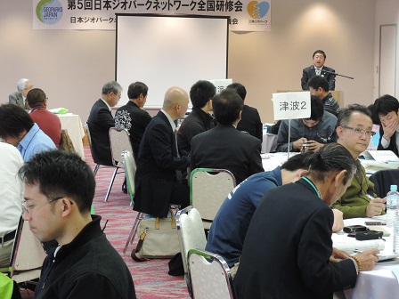 円卓の席に座る研修会の参加者たちと、スクリーンの横に立って話す市長の写真