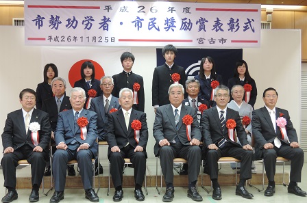 「市勢功労者・市民奨励賞表彰式」と書かれた横断幕の下で、椅子に座る10名の関係者と、後方に立つ学生6名の記念写真