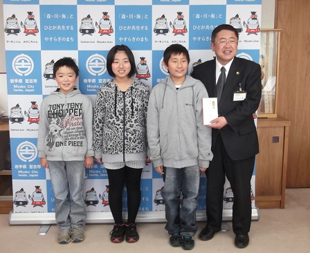 バックパネルの前で封筒を持つ市長と3名の小学生が並んでいる記念写真