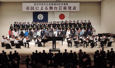 「市民による舞台芸術発表」と書かれた横断幕が掲げられた舞台で、ピアノやバイオリンなどの楽器を弾く方たちと、台の上に立ち歌う子供たちの写真
