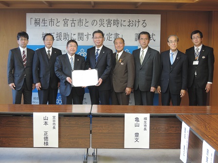 協定書を一緒に持つ市長と男性、関係者の男性6名が横一列に並んでいる記念写真