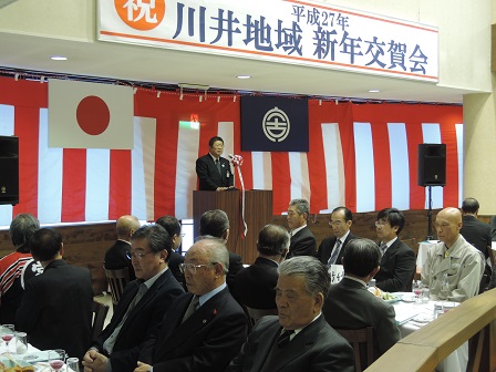 紅白の幕を張り、川井地域新年交賀会と書かれた横断幕の下であいさつをしている山本市長と、 座って聞いている参加者の方々の写真