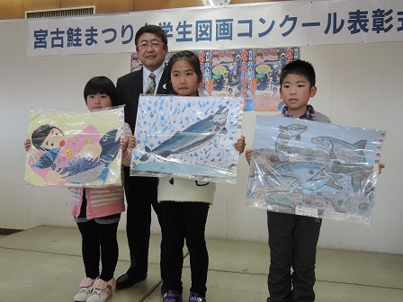 山本市長の前に図画コンクールで入賞した児童3人が、それぞれ描いた絵を持って並んで立っている写真