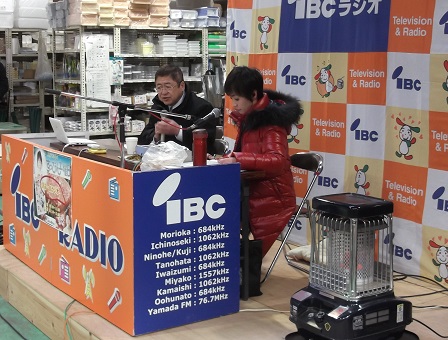 黒のジャンパーを着た市長が赤のフード付きのダウンコートを着た女性アナウンサーとストーブが置かれたIBCラジオの中継場所に座って番組に出演している写真