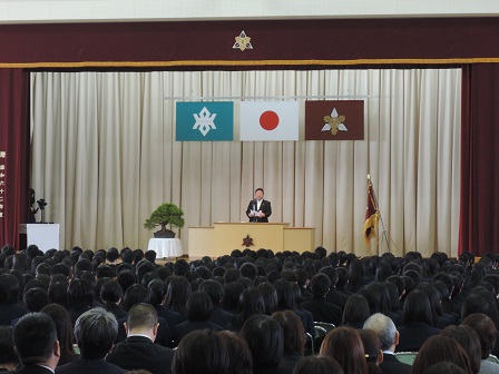 校旗と松の盆栽が置かれたステージ上の演台で祝辞を述べている市長と、座って聞いている卒業生と父兄の方々の写真