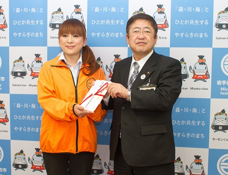 オレンジ色のウインドブレーカーを着た道の駅豊北のスタッフ菊地裕子さんが 市長へ寄附金の目録を渡している写真