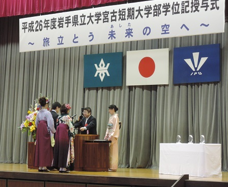 平成26年度岩手県立大学宮古短期大学部学位記授与式と書かれた横断幕がある ステージ上でスーツ姿の男性1人、袴姿の女性2人が市長より卒業証書を授与されている写真