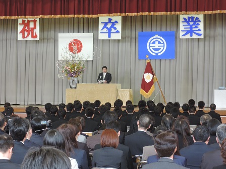 一文字ずつ祝卒業と書かれた幕と校旗が置いてあるステージ上で、祝辞を述べている市長と座って聞いている卒業生と父兄の方々の写真